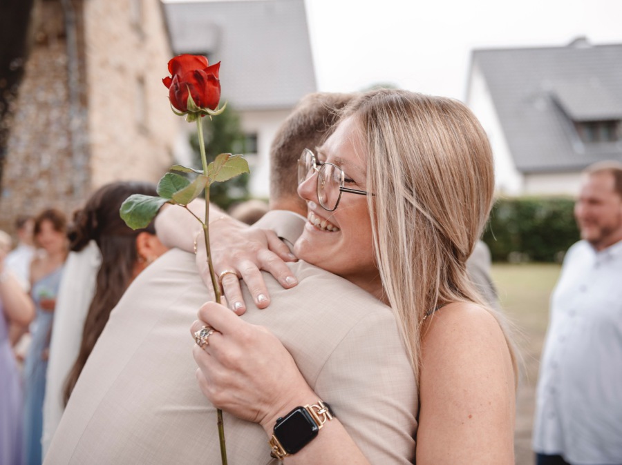Emotionale Umarmung beim Hochzeitsempfang – Hochzeitsgast gratuliert dem Bräutigam mit roter Rose